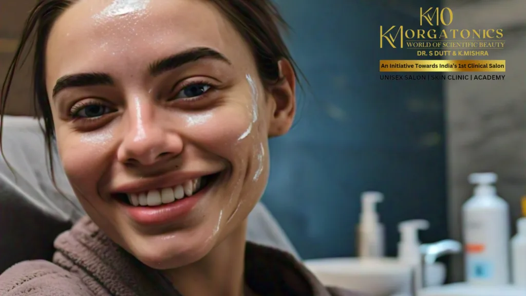 A woman with a bright smile and a skincare product applied to her face at KM Orgatonics. She's wearing a comfortable robe, suggesting a relaxing salon or spa environment.